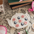 Teapot-shaped tic-tac-toe game with floral designs on a lace tablecloth