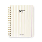A 2027 spiral-bound planner by Kate Spade New York, featuring a cream cover with thin vertical gold and pink stripes, gold wire binding, and black text.
