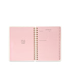 An open spiral-bound planner in pastel pink, showing a January 2023 weekly schedule page with lined sections and a left page that reads 'OFF TO A GOOD START'.
