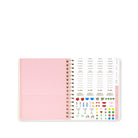 An open spiral-bound planner or agenda with a pale pink cover, showing printed weekly planner pages and small colorful stickers along the right side on a white background.