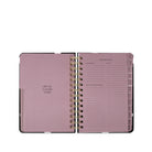 An open spiral-bound planner with dusty pink pages and gold-toned metal rings, shown against a plain white background.