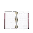 An open spiral-bound planner showing a monthly calendar layout in white pages with dusty pink sidebar accents and gold-toned wire binding, photographed on a clean white background.