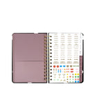 An open spiral-bound planner or agenda with a dusty mauve-pink cover on the left and a white interior page filled with small printed labels, icons, and colored stickers on the right.