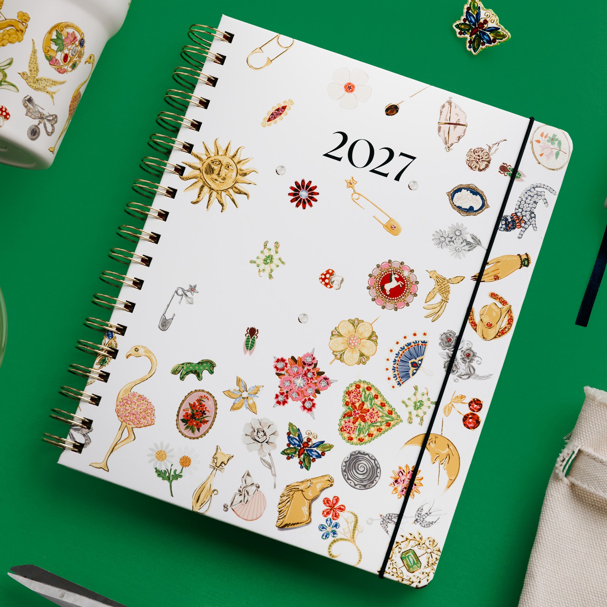 A 2027 spiral-bound planner with a white cover decorated with colorful vintage-style fashion illustrations, shown on a bright green background with styled props.