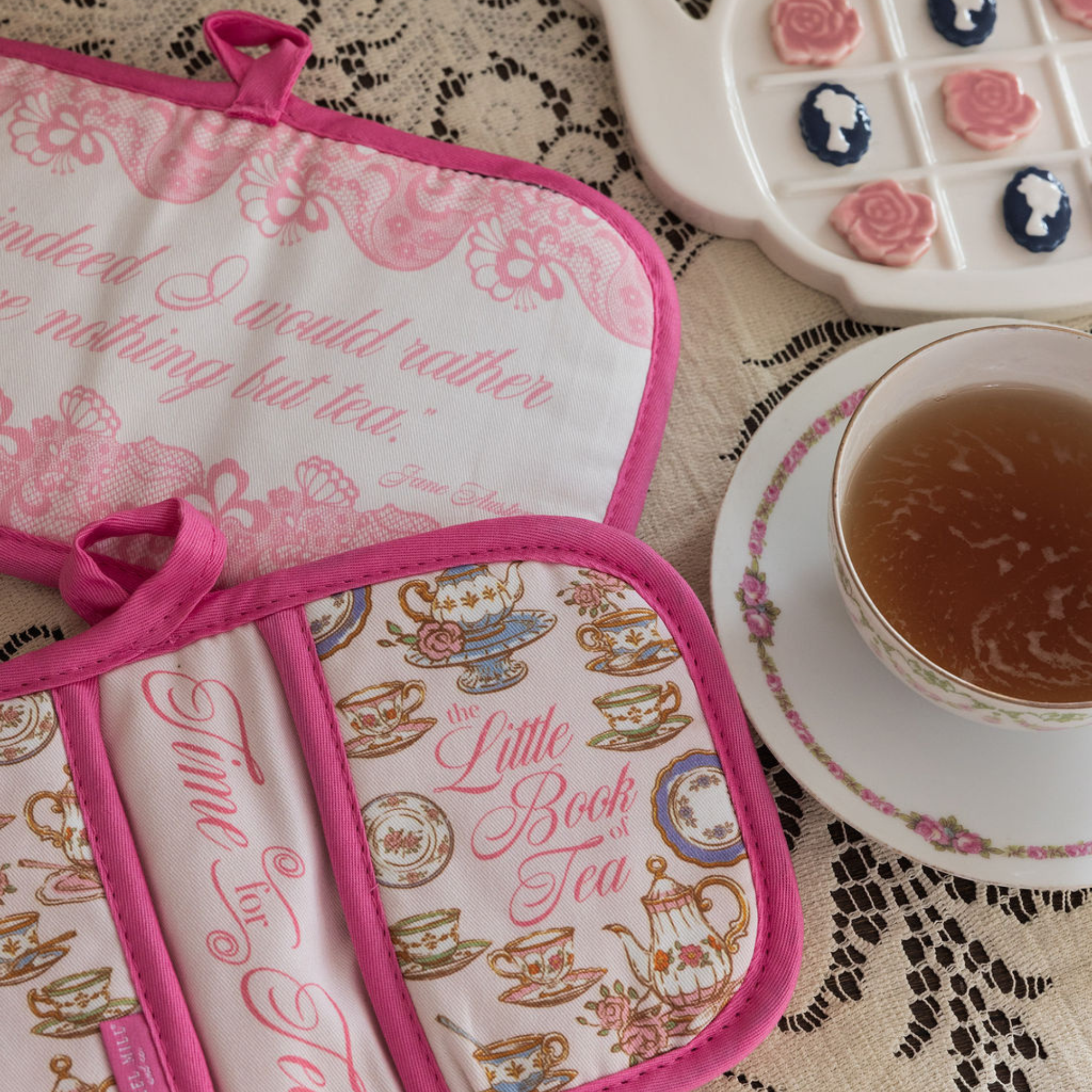 Tea-themed pot holders with decorative text and designs on a lace tablecloth with a cup of tea.
