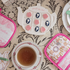 Tea-themed kitchen accessories including a teapot-shaped cookie cutter, tea cup, and pot holders on a lace tablecloth.