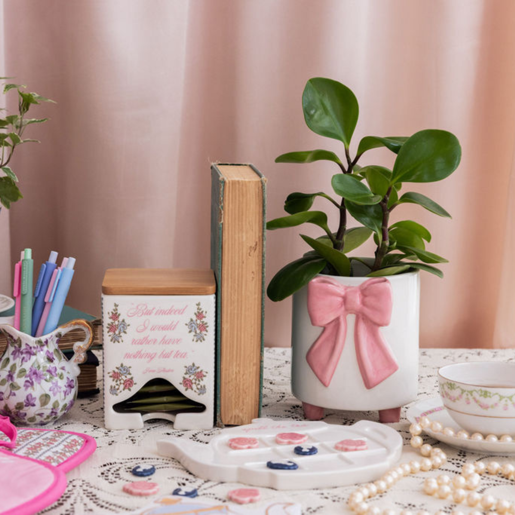 Decorative items including a potted plant with a pink bow, books, and a teapot on a table with a pink curtain background.