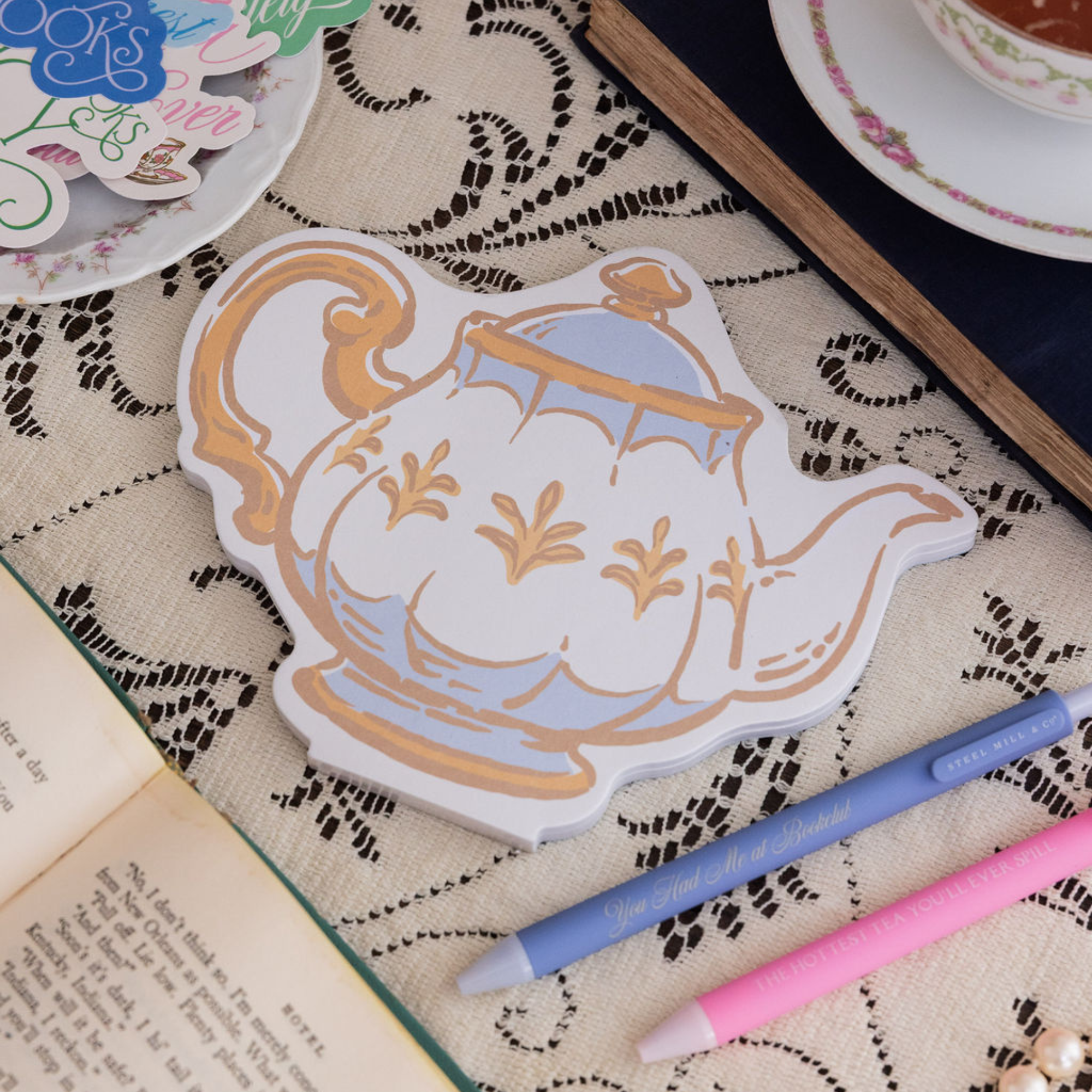 Decorative teapot-shaped notepad on a textured surface with pens and books.