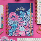 A Lilly Pulitzer 2026 daily planner with a navy blue cover, featuring a vibrant pink and blue floral pattern and gold foil lettering, styled on a bright pink desk with a keyboard, sticky notes, and other stationery.