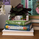 Stack of books with decorative covers on a white surface, surrounded by flowers and plants.