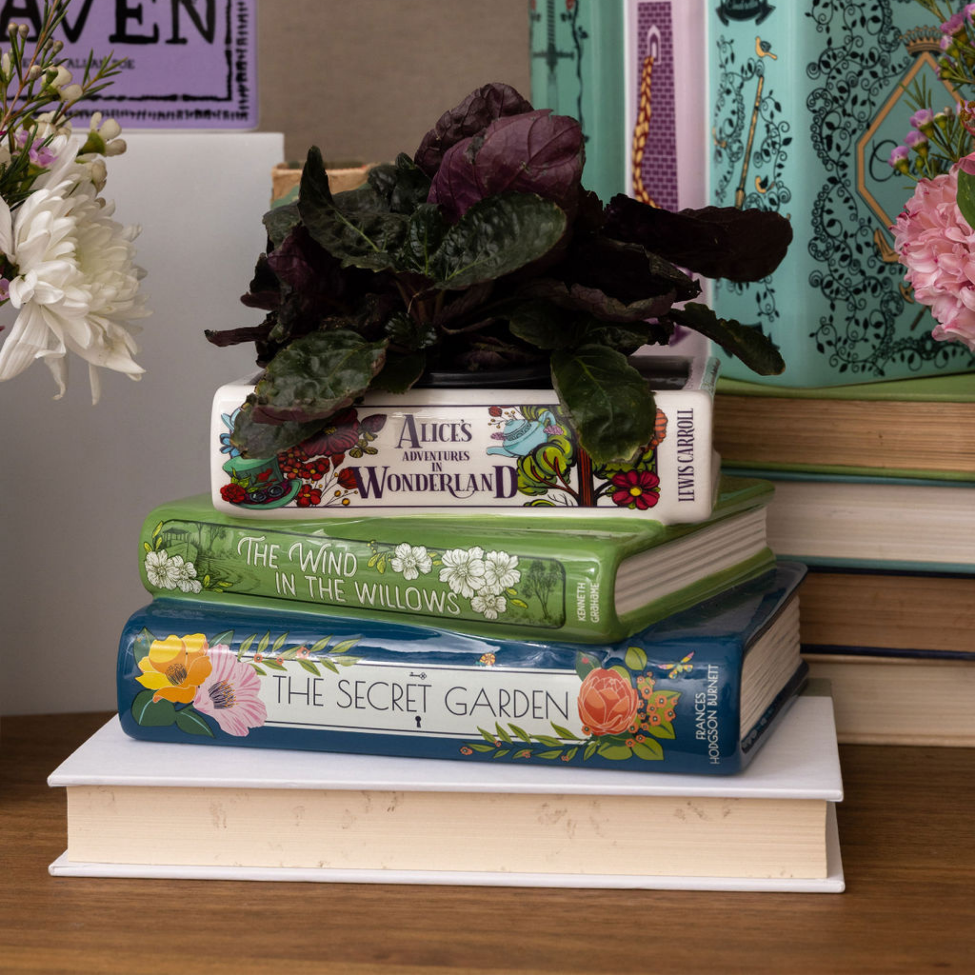 Stack of books with decorative covers on a white surface, surrounded by flowers and plants.