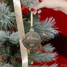 A close-up of a hand with red nail polish hanging suriel trading co. embossed gold ornament with gold hanging ribbon from a frosted blue-green pine tree with a cream ribbon and warm string lights against a red background.