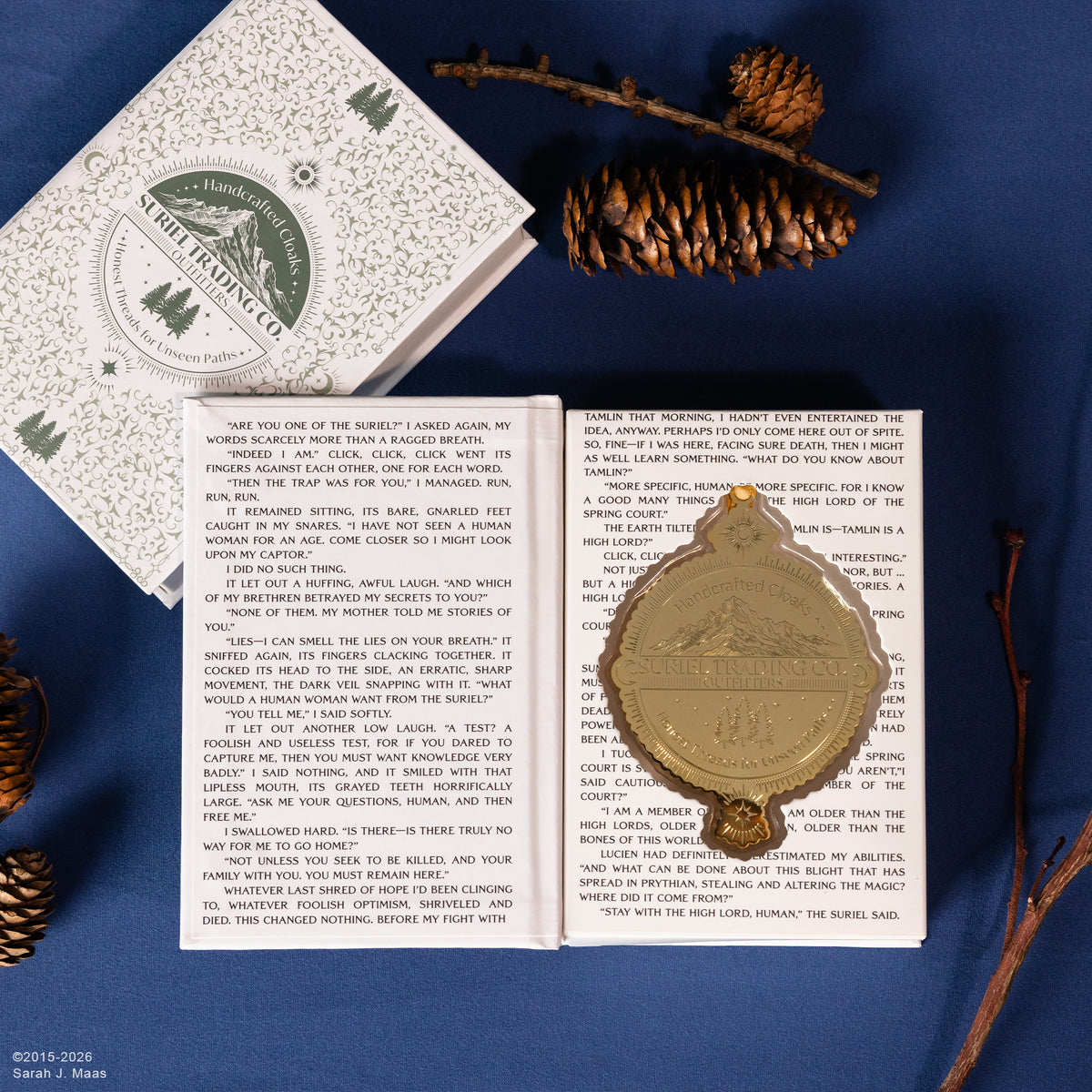 An open vintage book spread on a dark blue fabric background, featuring a suriel trading co. embossed gold ornament with gold hanging ribbon resting on the right page with book packaging next to it