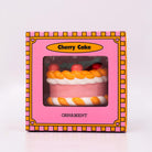 Packaged ornament cake with pink filling, orange and white striped sides, and red cherries on a white background