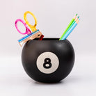 Black billiard ball-shaped holder with number 8 holding stationery items on a white background
