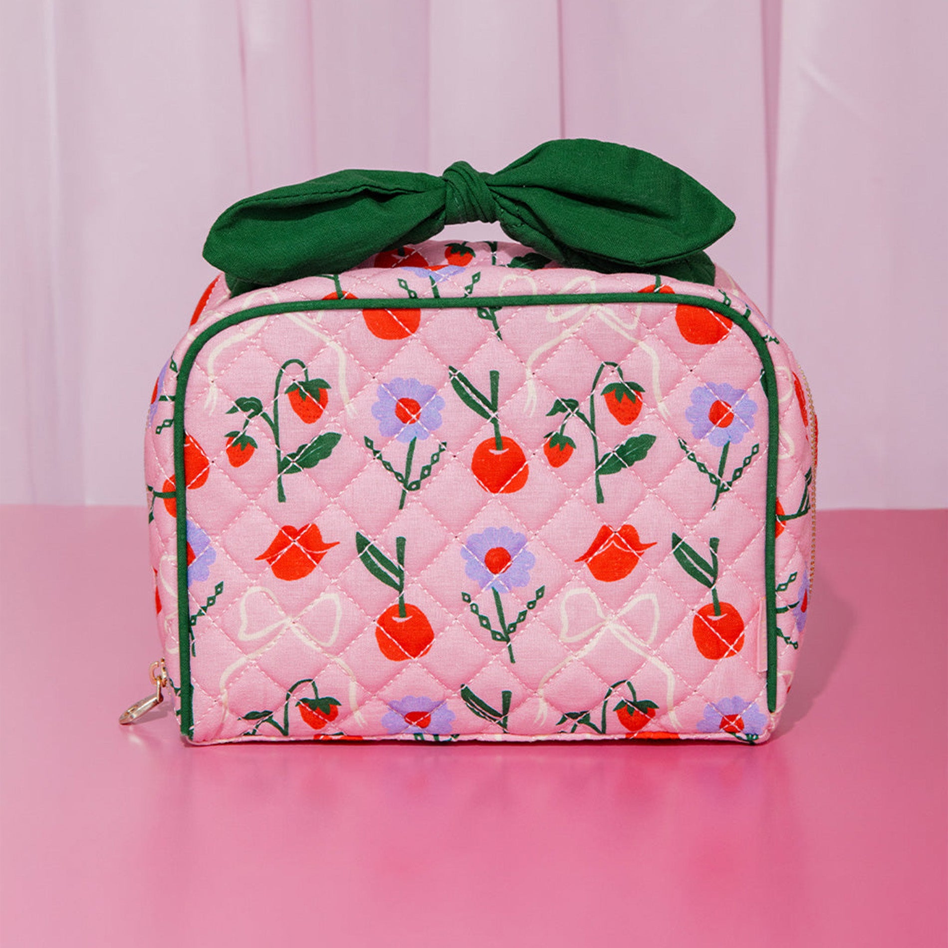 Pink quilted bag with floral and fruit patterns and a green bow on a pink background