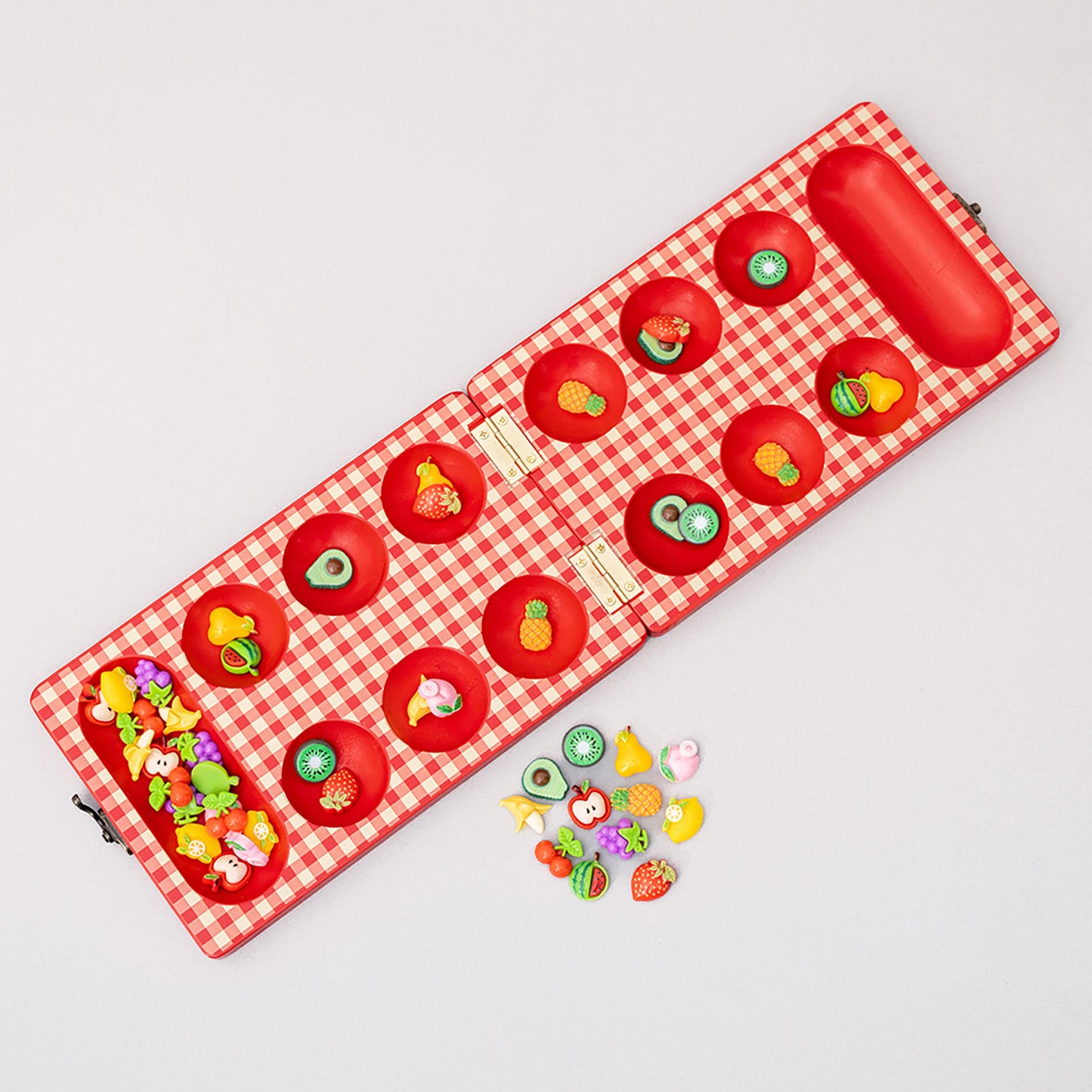 Red checkered mancala board with fruit-themed items on a light gray background