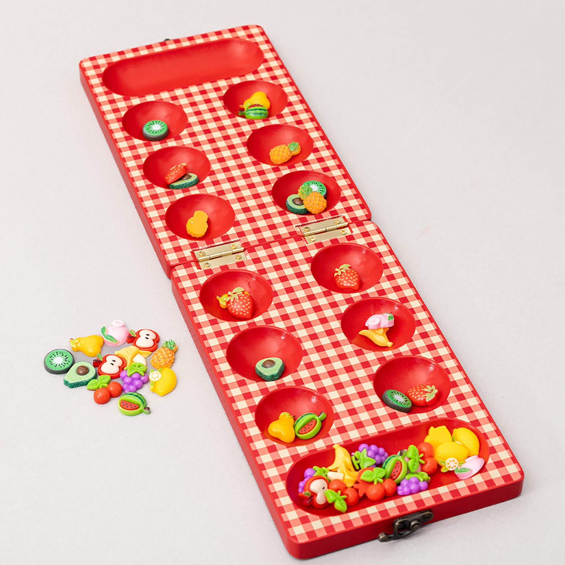 Red checkered mancala board with fruit-themed items on a light gray background
