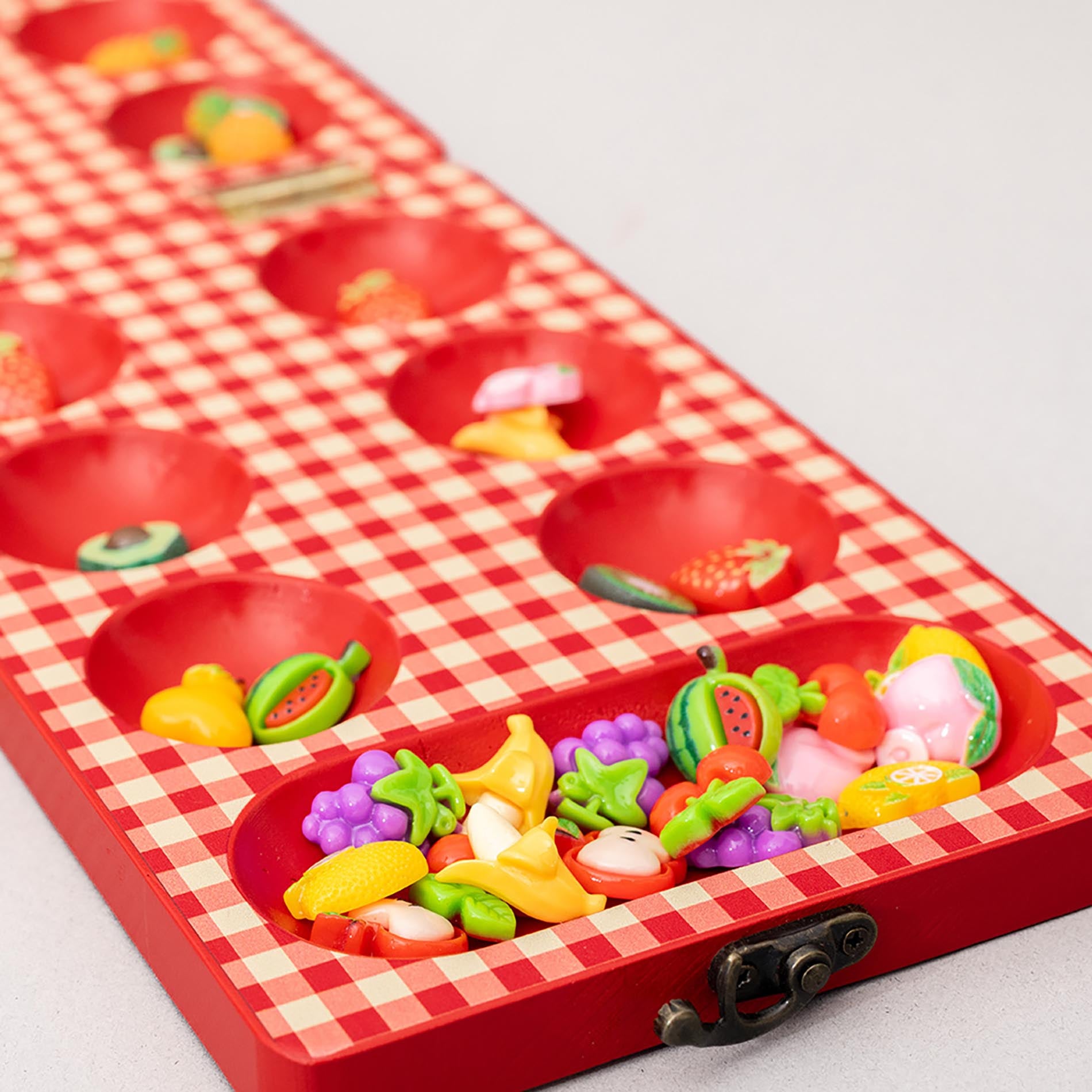Up close of red checkered mancala board with fruit-themed items on a light gray background