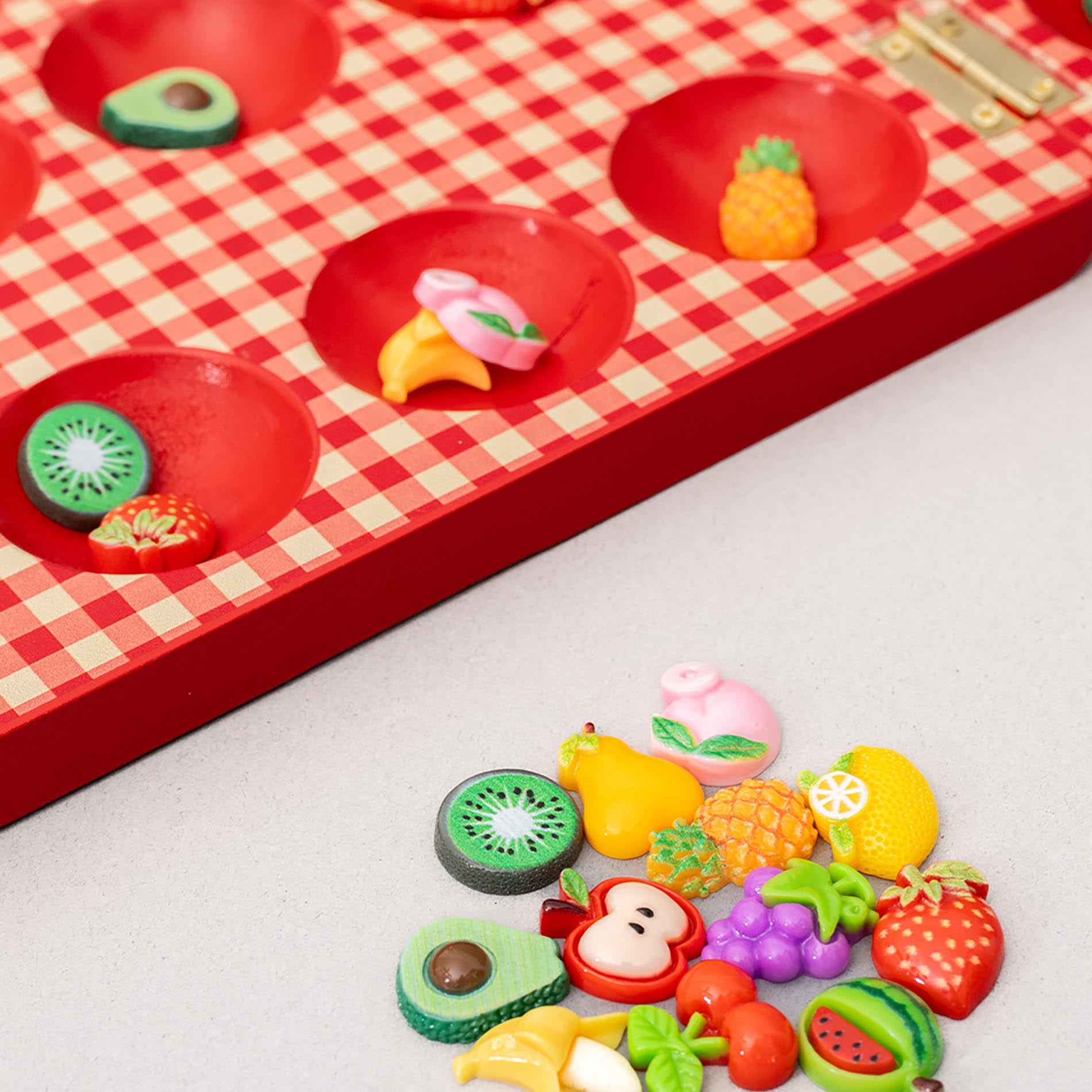 Up close of pieces of red checkered mancala board with fruit-themed items on a light gray background