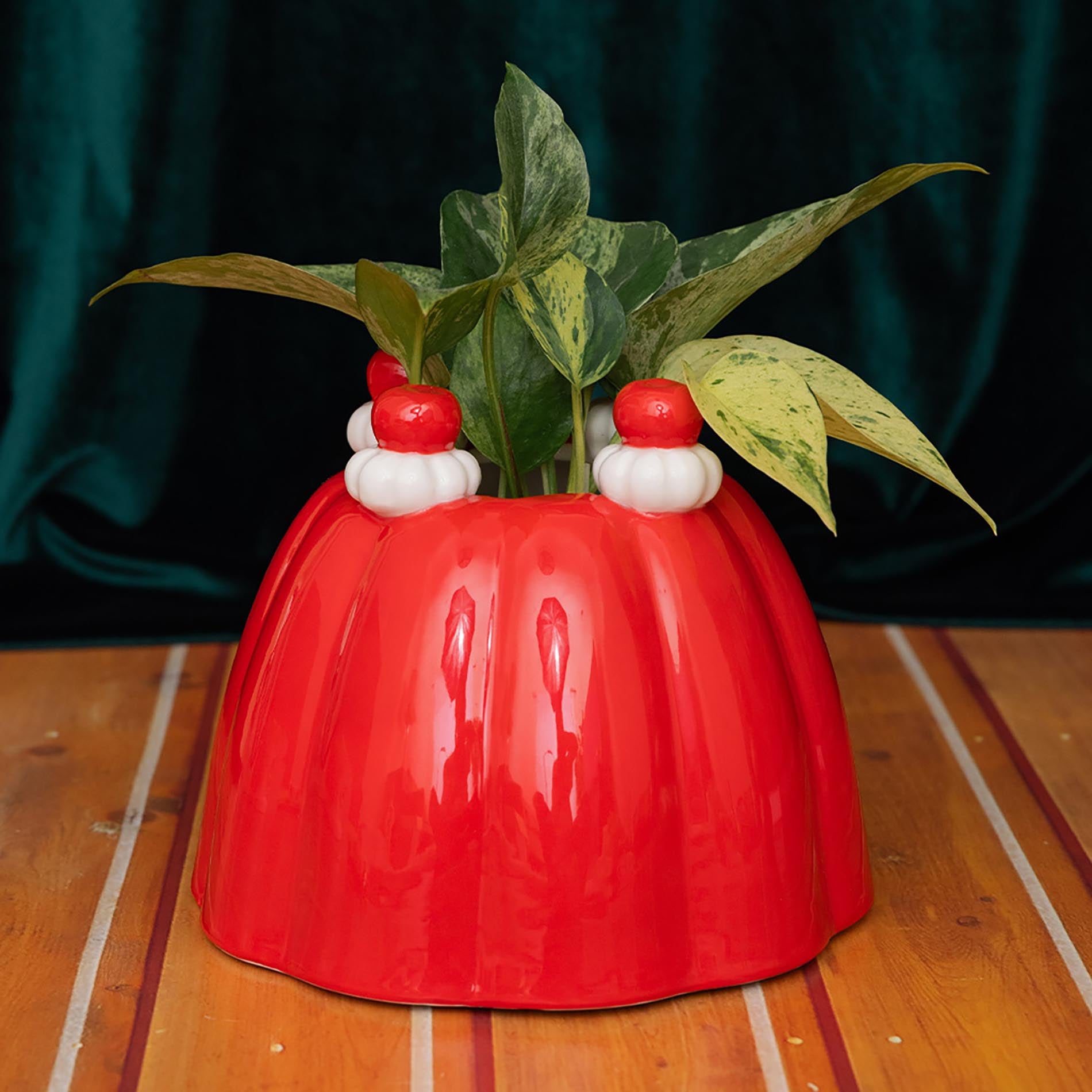 Red jelly vase planter with small red and white figures on top, containing a green plant, on a wooden surface.