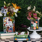 Books with floral designs on covers displayed against a green bush background.