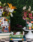 Books with floral designs on covers displayed against a green bush background.