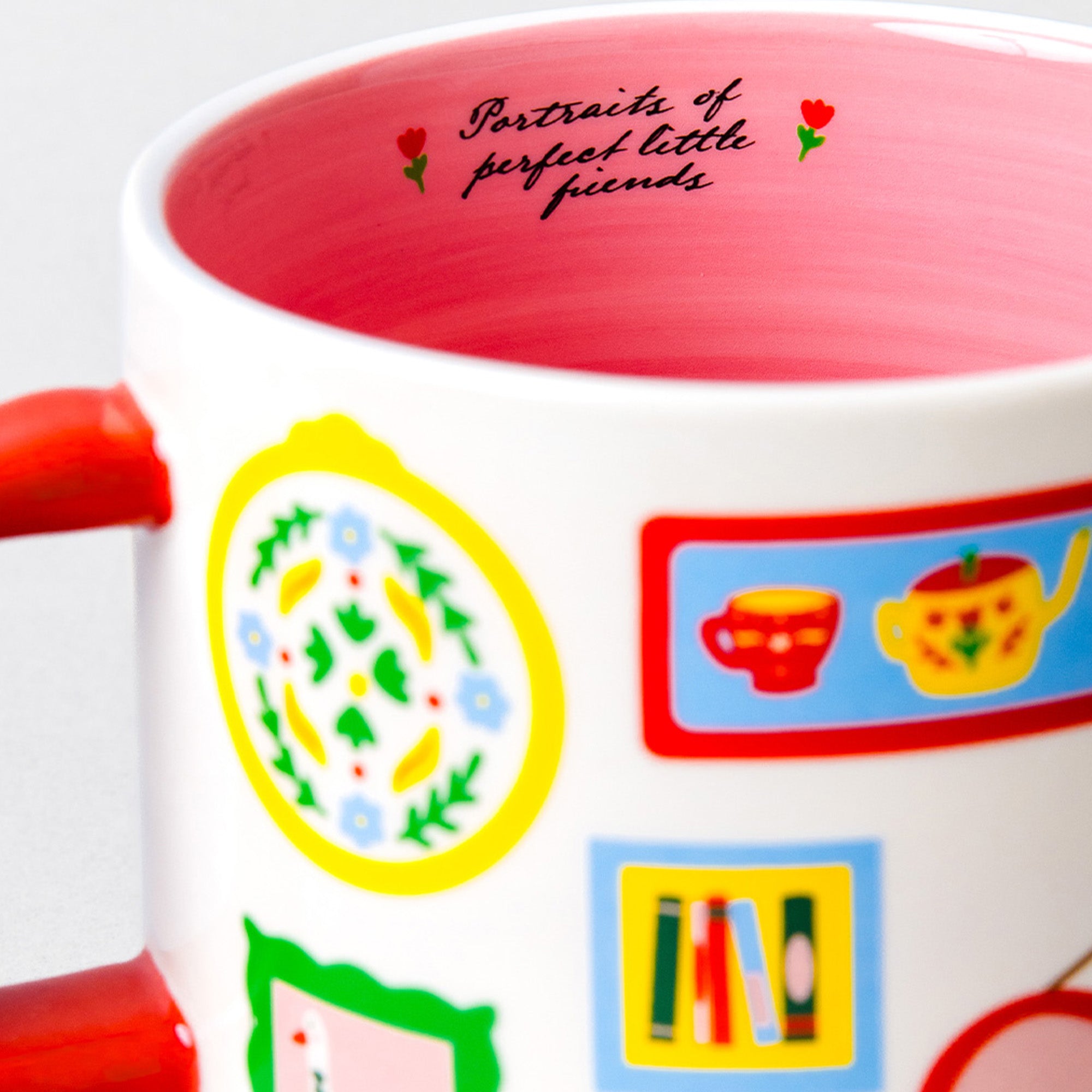 A close-up of a white ceramic mug with a red handle and red interior, decorated with colorful illustrated motifs and the phrase 'Portraits of perfect little friends' printed inside.