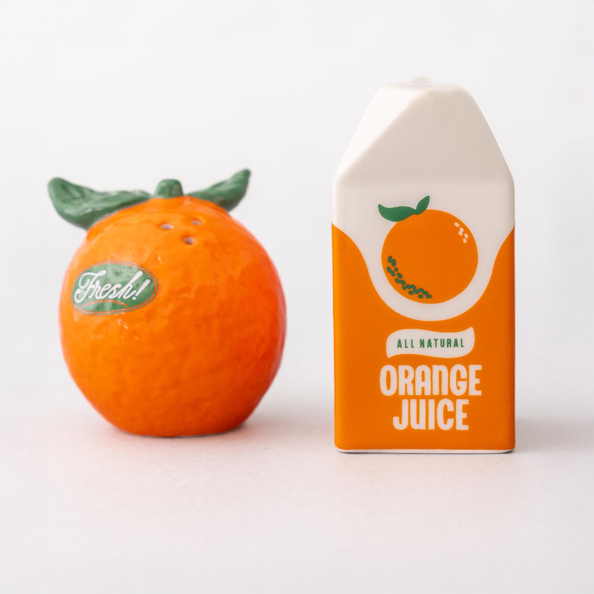 Decorative salt and pepper shaker shaped like oranges next to a box of all natural orange juice on a white background
