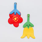 Two flower-shaped luggage tags on a light gray background. One tag is red with a blue top and yellow center, and the other is yellow with a green top and yellow center.