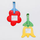 Two flower-shaped luggage tags on a light gray background. One tag is red with a blue strap and a white insert, and the other is yellow with a green strap and a white insert.