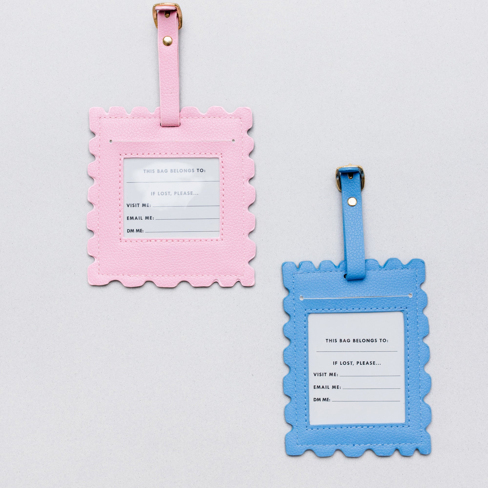 Two scalloped luggage tags on a light gray background, one in pink and one in blue, each with a white insert card and gold-tone metal hardware.
