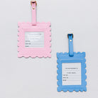 Two scalloped luggage tags on a light gray background, one in pink and one in blue, each with a white insert card and gold-tone metal hardware.