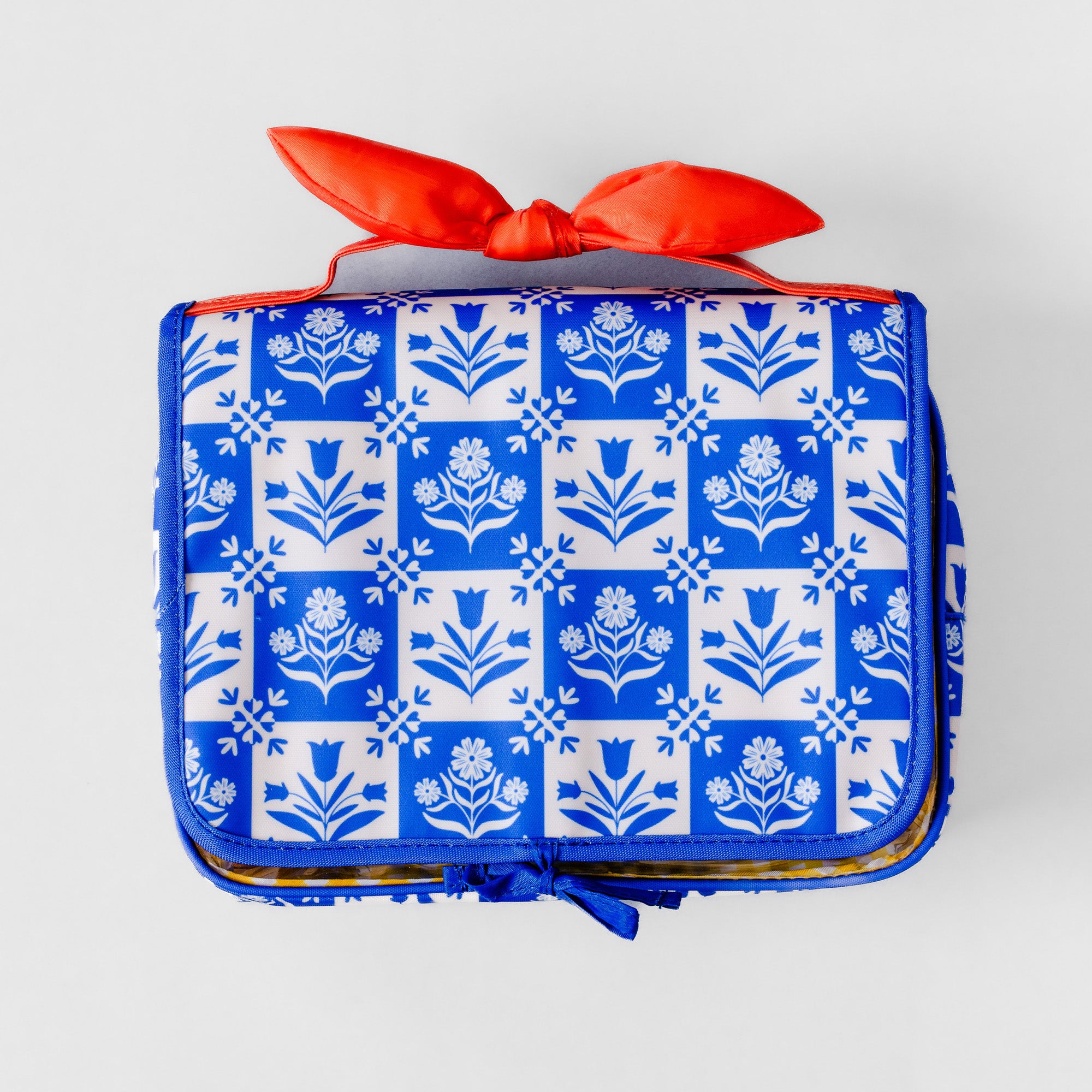 A rectangular zippered travel or toiletry bag with a blue and white floral tile pattern, featuring a bright orange-red satin ribbon tied in a bow on top, photographed against a light gray background.
