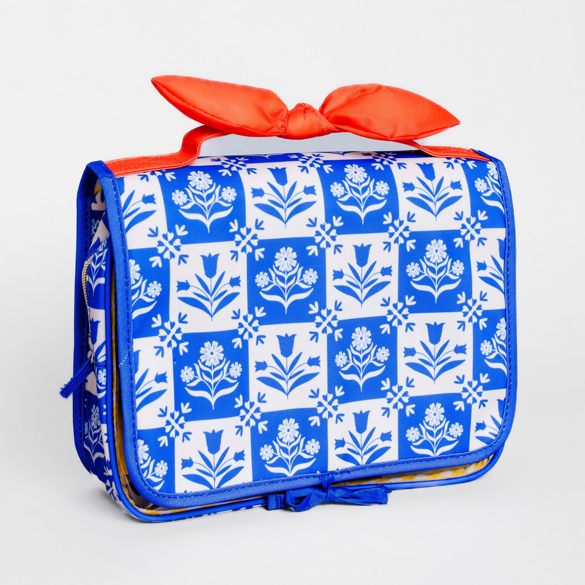 A rectangular soft-sided bag with a blue and white floral block-print pattern, featuring a bright orange-red satin bow on top and a matching blue zipper pull, shown on a light gray background.