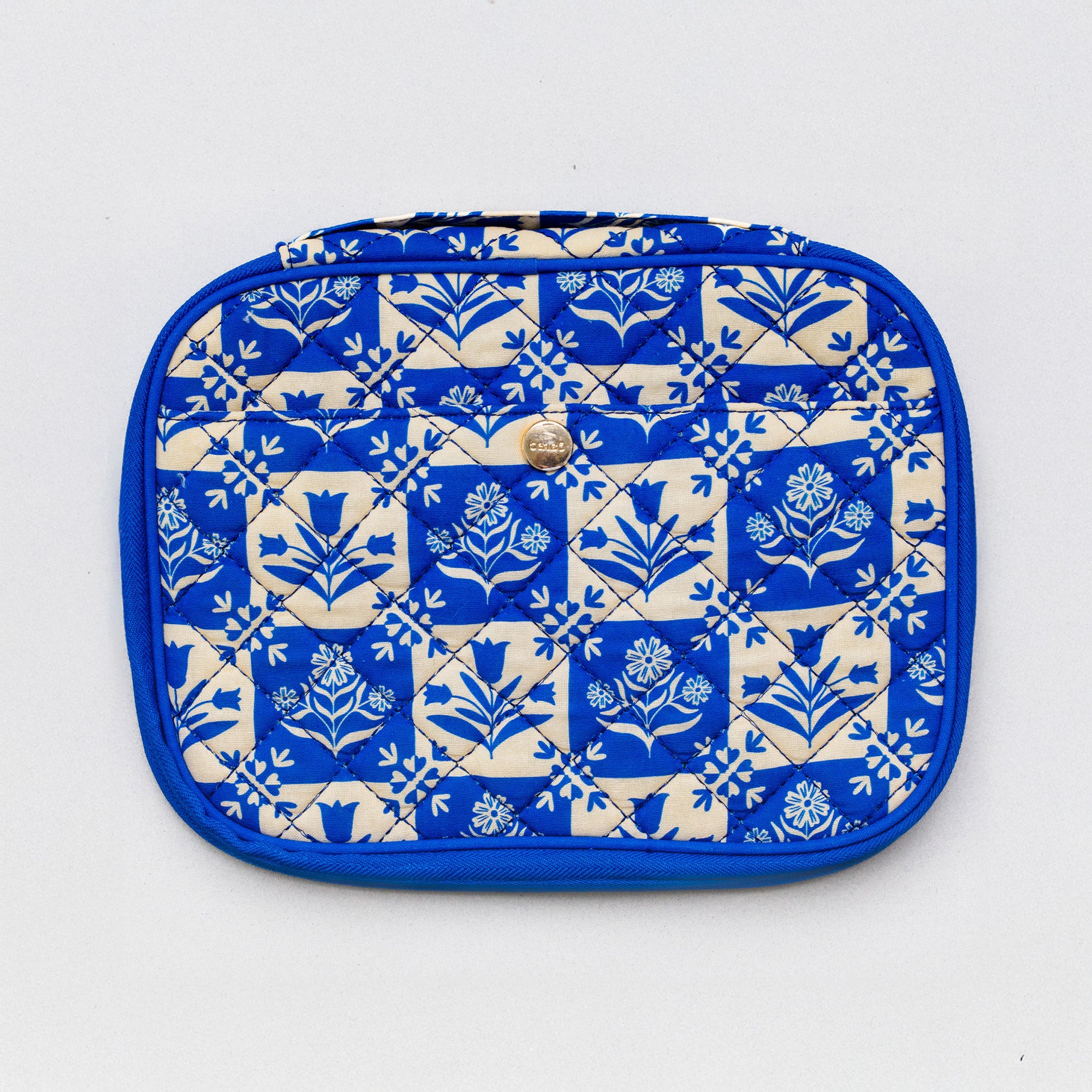 A quilted cosmetic pouch with a blue and cream floral block-print pattern, featuring a small gold metal logo on the front.