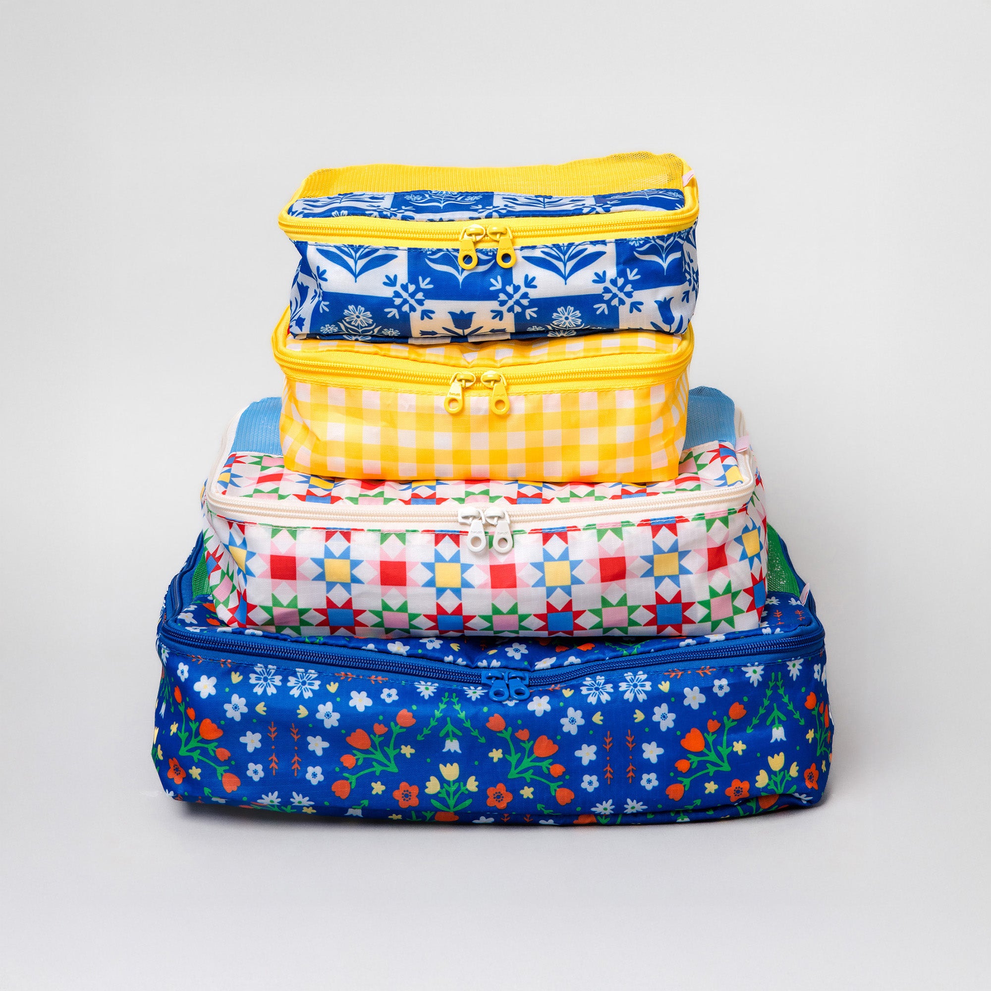 A stack of four colorful zippered fabric storage bags with various patterns including gingham, floral, and geometric, set against a plain light gray background.