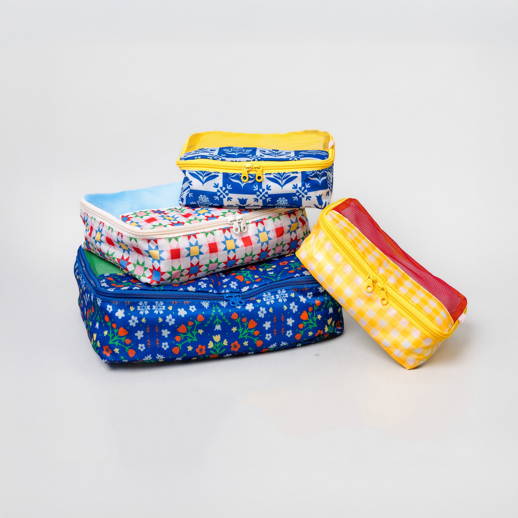 Four fabric zippered pouches stacked and leaning on a light gray studio background. The set includes blue floral, red-and-white gingham, yellow-and-white gingham, and blue-and-white patterned pouches with yellow and red zippers.