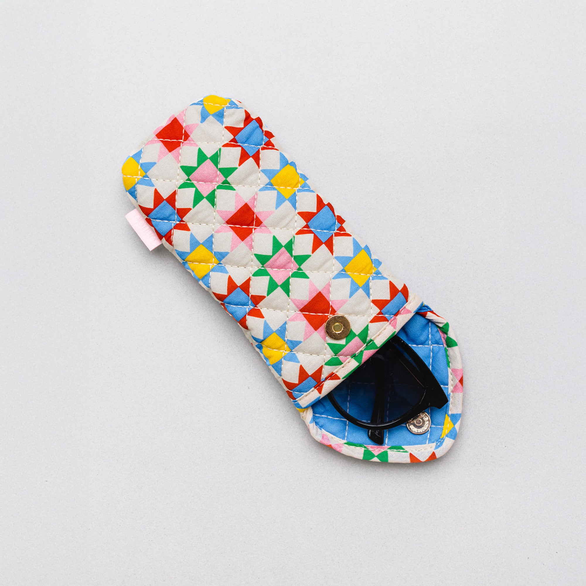 A quilted fabric pouch with a colorful geometric star pattern in red, yellow, green, blue, and pink on a white background, shown with a pair of sunglasses inside on a light gray background.