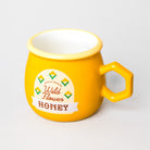 A bright yellow ceramic honey mug with a hexagonal handle, featuring a white and brown 'Wild Flower Honey' label on the front, set against a neutral light gray background.