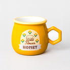 A bright yellow ceramic mug with a hexagonal handle, featuring a printed label that reads 'Wild Flower HONEY' with small flower illustrations, on a light gray background.
