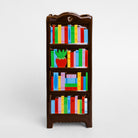 A small dark brown bookshelf vase decorated with colorful book spines and a red potted plant on the middle shelf, shown against a plain light gray background.