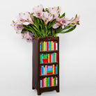 A decorative dark brown bookshelf vase filled with pink alstroemeria flowers, set against a plain light gray background.