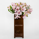 A tall, narrow glossy brown ceramic vase with a gold heart logo reading 'BOOMZ', filled with a bouquet of light pink alstroemeria flowers, set against a plain white background.