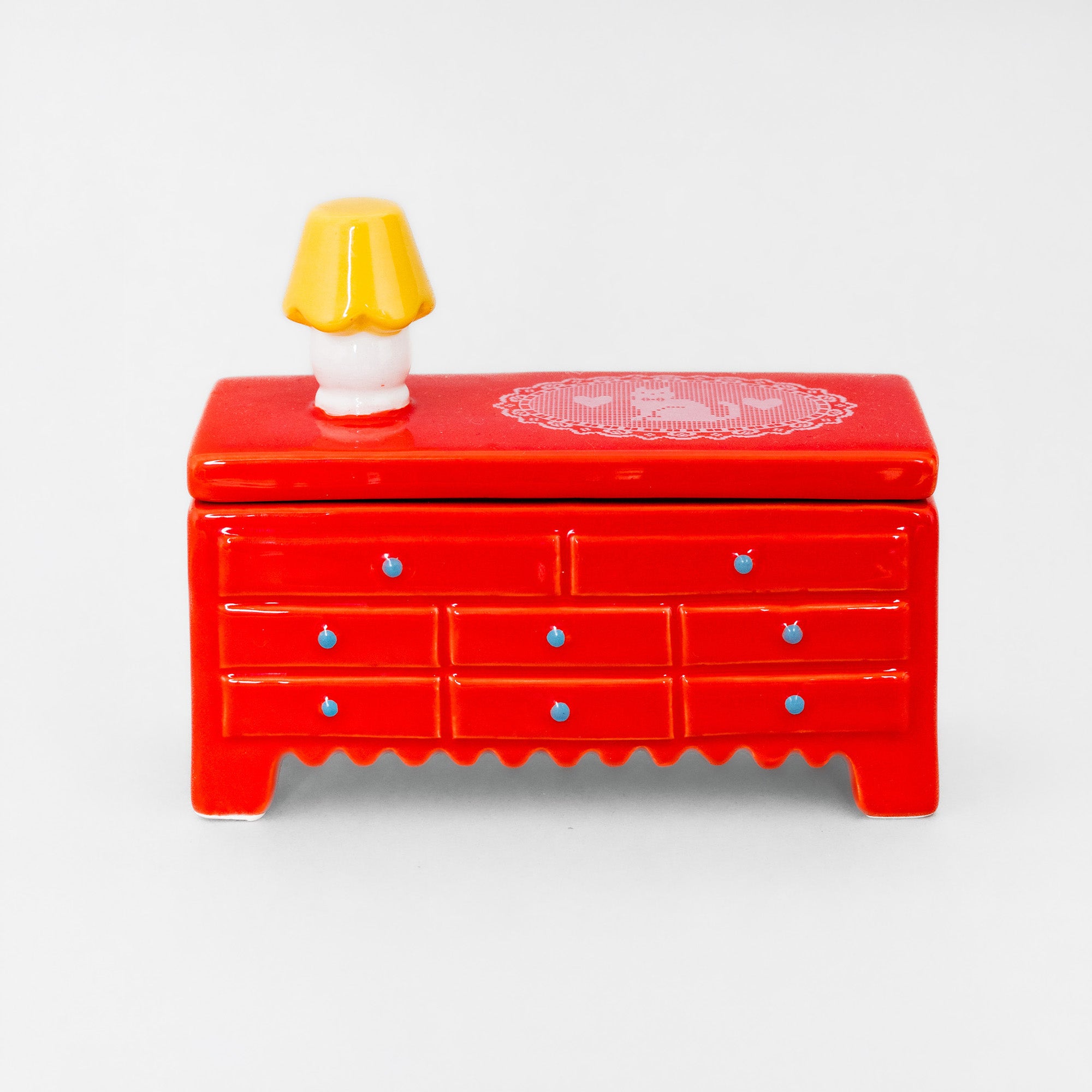 A small red ceramic jewelry box with multiple drawers, blue knob details, a scalloped front edge, and a tiny yellow-and-white lamp on top, photographed on a plain light gray background.