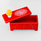 A red rectangular jewelry box with a hinged lid, shown open on a light gray background. The lid features a white printed lace-like oval design with a cat motif and a small yellow-and-white mushroom-shaped charm resting on the corner.