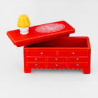 A miniature red dresser with drawers and a matching lid, featuring a small yellow and white lamp on top, set against a plain light gray background.