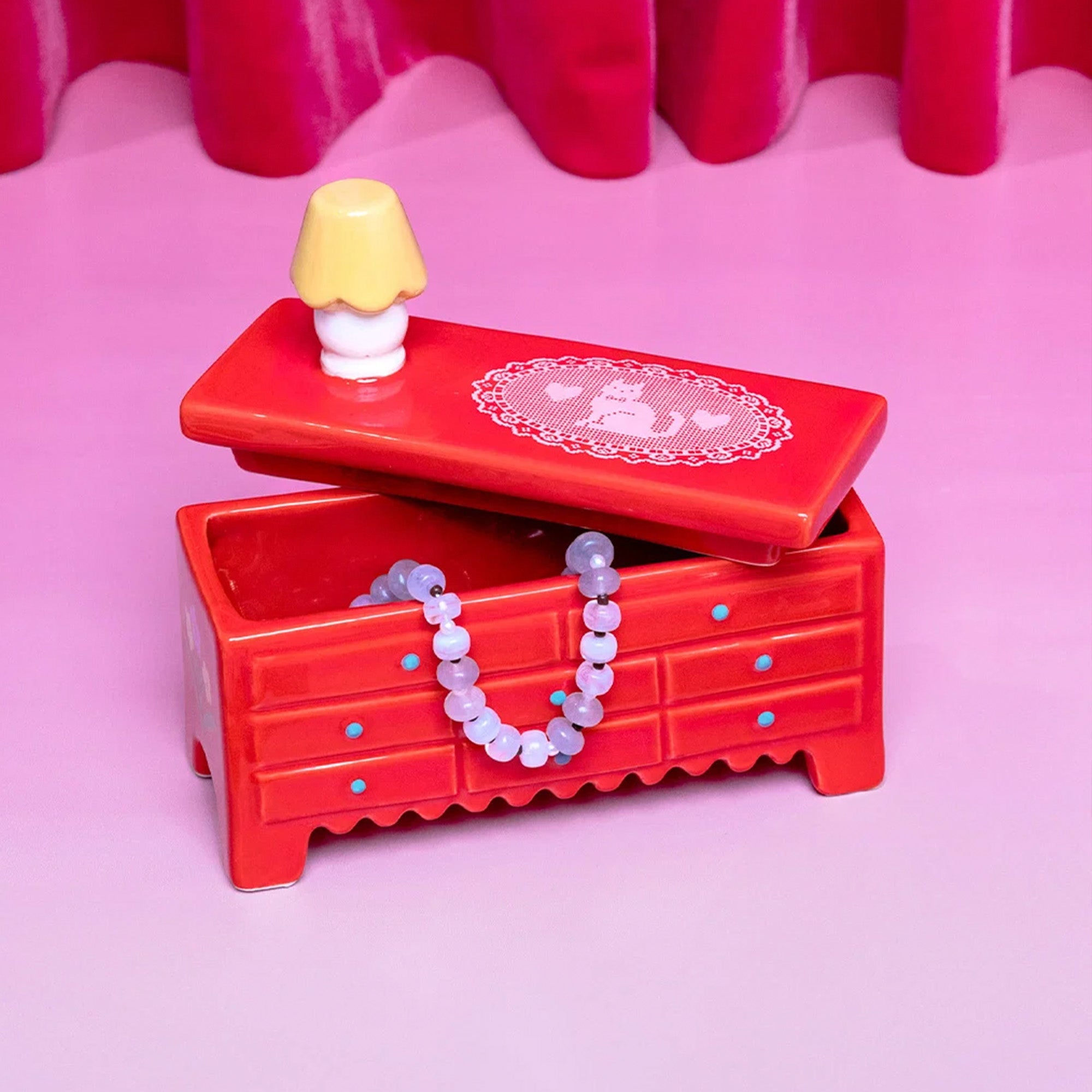 A red ceramic jewelry chest with a matching red lid, decorated with a white lace-like pattern and a small yellow-and-white lamp figurine on top. A beaded lavender necklace is draped over the front, and the chest sits on a pink surface with a pink background and red draped fabric behind it.