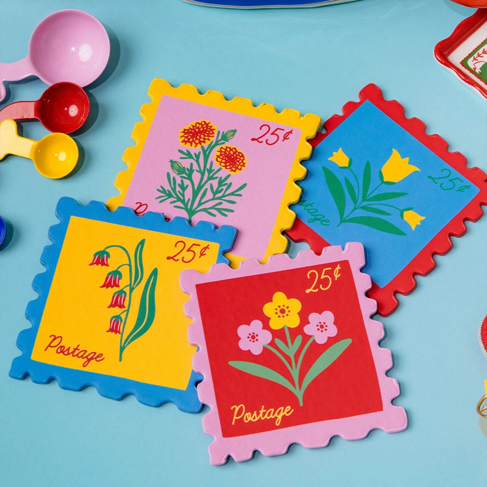 Four colorful postage-stamp-shaped coasters with scalloped edges, each printed with a different flower illustration and the word "Postage" plus a 25¢ symbol, arranged on a light blue surface with small toy measuring cups partly visible at the left edge.