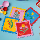 Four colorful postage-stamp-shaped coasters with scalloped edges, each printed with a different flower illustration and the word "Postage" plus a 25¢ symbol, arranged on a light blue surface with small toy measuring cups partly visible at the left edge.